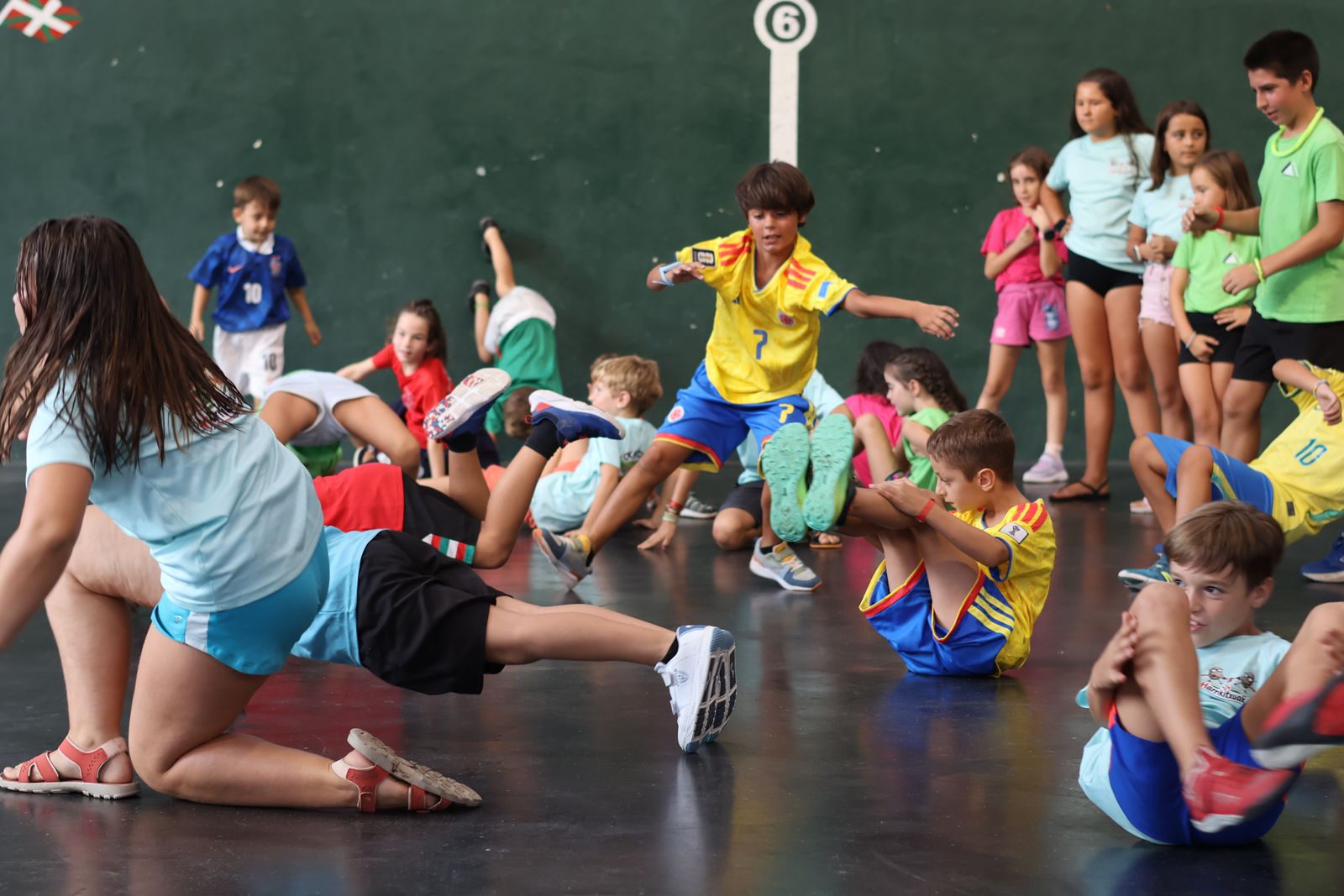 Grupo grande de niños haciendo el calentamiento sentados en el suelo del frontón de Okondo